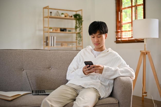 Giovane asiatico rilassato che usa il suo smartphone mentre si rilassa sul divano nel suo salotto