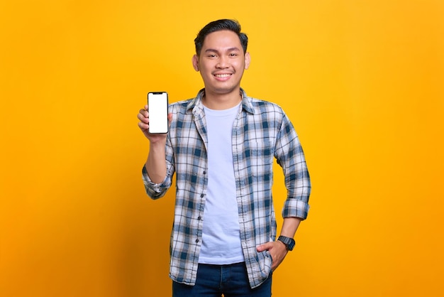 Giovane asiatico sorridente in camicia a quadri che mostra lo schermo vuoto del telefono cellulare che consiglia l'app isolata su sfondo giallo
