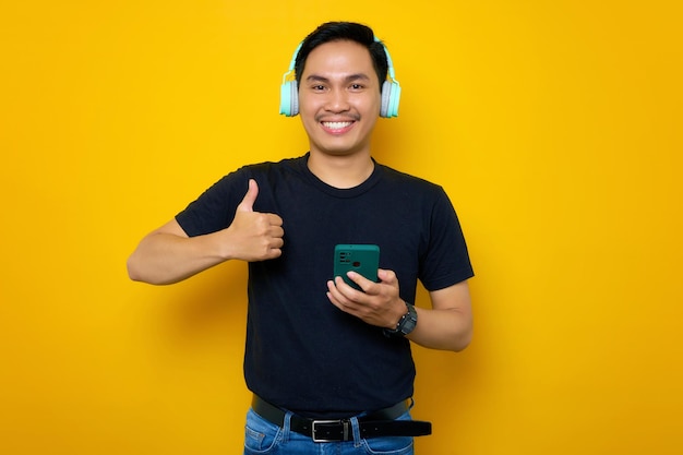 Giovane asiatico sorridente in maglietta casual con telefono cellulare e cuffie wireless che mostrano i pollici in su isolati su sfondo giallo Concetto di stile di vita delle emozioni delle persone