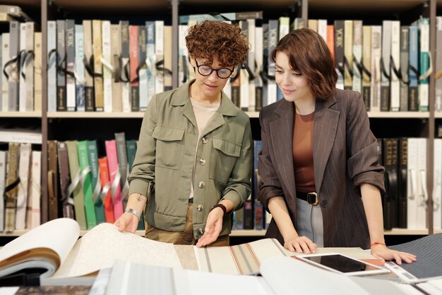 Giovane assistente di negozio femminile bruna fiduciosa che aiuta il cliente a scegliere un campione di nuovi sfondi in studio contemporaneo di interior design