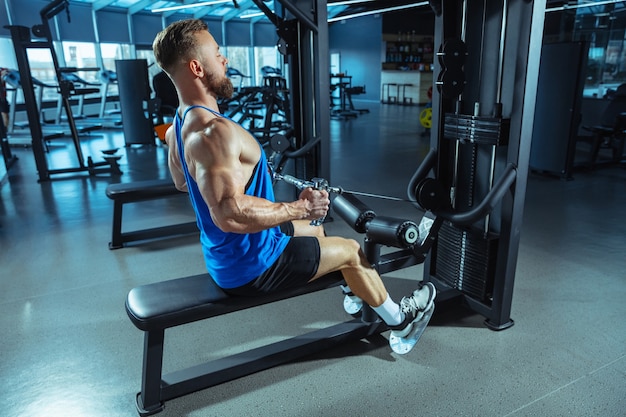 Giovane atleta caucasico muscoloso che si allena in palestra, fa esercizi di forza, pratica, lavora sulla parte superiore del corpo, tira su pesi e bilancieri. Fitness, benessere, concetto di stile di vita sano, lavoro.