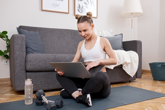 Giovane atleta femminile seduto a gambe incrociate sul tappetino vicino a bottiglia e manubri e laptop di navigazione durante l'allenamento fitness a casa