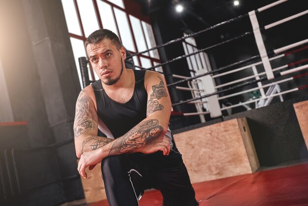 Giovane atleta muscolare concentrato in abbigliamento sportivo colorato che riscalda i suoi muscoli prima dell'allenamento nella palestra di boxe nera