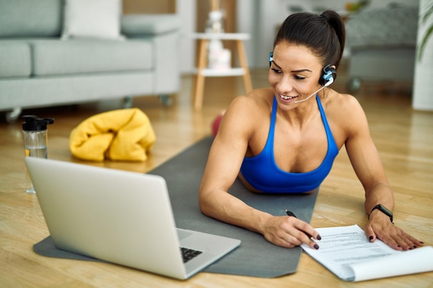 Giovane atleta sorridente che utilizza il computer e prende appunti mentre fa il suo piano di esercizi nel soggiorno