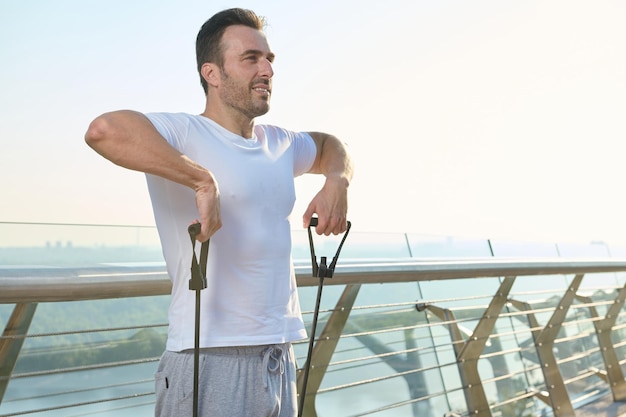 Giovane atleta sportivo europeo durante l'allenamento all'aperto con fascia di resistenza in gomma Atleta di mezza età che fa esercizi su spalle e braccia utilizzando un espansore elastico sul ponte di vetro della città all'alba