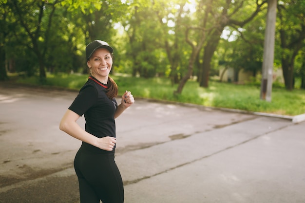 Giovane atletica sorridente bella ragazza bruna in uniforme nera e allenamento con berretto, facendo esercizi sportivi e correndo, guardando indietro sul sentiero nel parco cittadino all'aperto