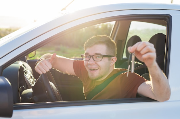 Giovane autista che mostra le chiavi della macchina e il pollice in alto felice. Uomo che tiene la chiave dell'auto per la nuova automobile. Auto a noleggio o concetto di patente di guida con guida maschile nella splendida natura durante un viaggio su strada.