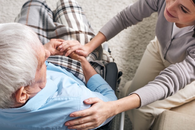 Giovane badante femminile solidale seduto da un uomo anziano in sedia a rotelle e tenendo la mano sulla sua spalla mentre lo conforta