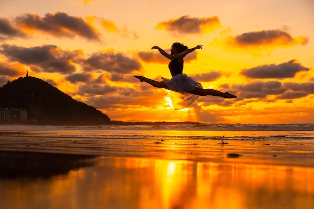 Giovane ballerina sulla spiaggia al tramonto che esegue un salto con il mare sullo sfondo con il sole riflesso nell'acqua