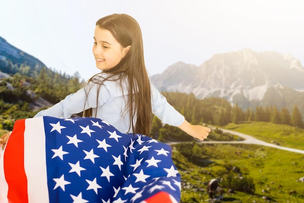 Giovane bambina che tiene la bandiera americana sullo sfondo della natura incredibile cielo, montagna e prato al tramonto.