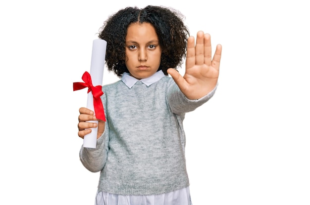 Giovane bambina con i capelli afro che tiene il diploma di laurea con la mano aperta che fa il segnale di stop con un gesto di difesa dell'espressione seria e sicura