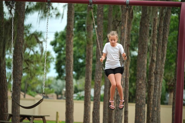 Giovane bambina felice che gioca da sola volando in alto sulle altalene durante il fine settimana estivo giornata di sole Sicurezza e ricreazione sul concetto di parco giochi