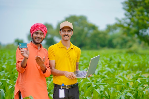 Giovane banchiere o agronomo indiano che mostra alcuni dettagli agli agricoltori in laptop nel campo agricolo.