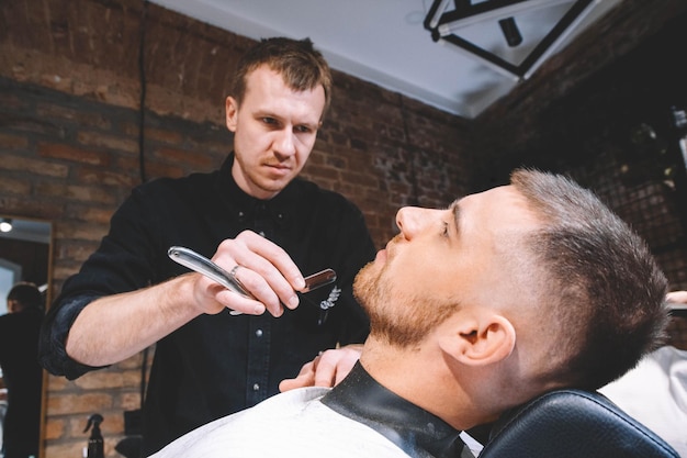 Giovane barbiere maschio fa un cliente barba taglio di capelli con rasoio vintage. Processo di rasatura della barba in Barberia