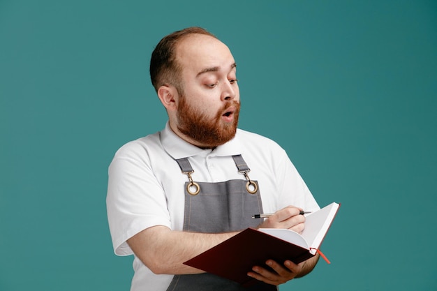 Giovane barbiere maschio sorpreso che indossa una camicia bianca e un grembiule da barbiere con penna e blocco note guardando il blocco note isolato su sfondo blu