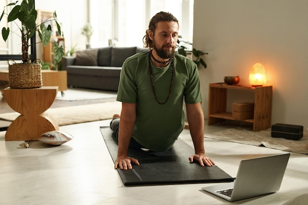 Giovane barbuto che guarda lezioni online sul laptop e fa esercizi di yoga sul tappetino per esercizi a cupola