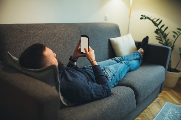 Giovane barbuto che si appoggia sul divano e guardando lo schermo dello smartphone a casa sua dopo il lavoro.