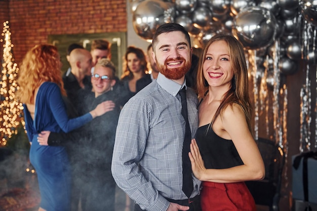 Giovane barbuto con la sua ragazza in piedi insieme contro i loro amici nella stanza decorata di natale e festeggia il nuovo anno.
