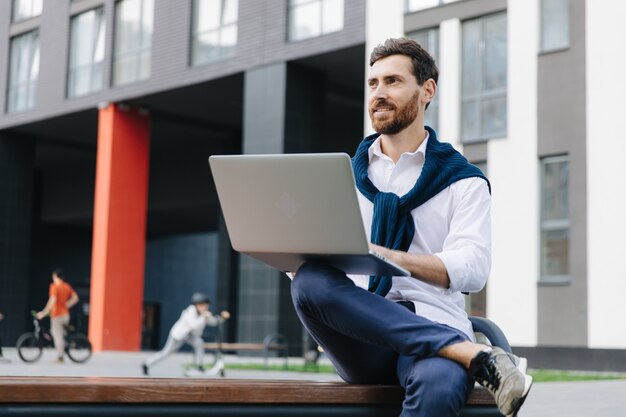 Giovane barbuto in camicia bianca e jeans seduto vicino a un edificio moderno e che si gode il lavoro a distanza su un laptop wireless. Concetto di persone e tecnologia.
