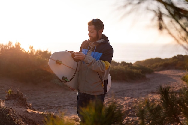 Giovane barbuto in giacca che cammina al tramonto sulle dune della spiaggia con la sua tavola da surf sotto il braccio il tempo libero e gli hobby concetto copia spazio per il testo