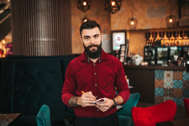 Giovane barista barbuto bello in camicia rossa che lavora nel moderno bar caffetteria.