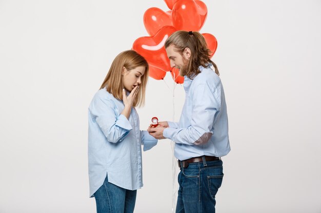 Giovane bel ragazzo dà un anello a una ragazza su uno sfondo bianco con palloncino rosso cuore