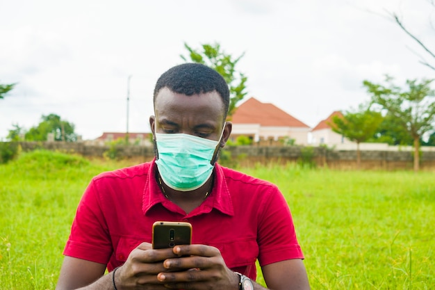 Giovane bell'uomo africano seduto fuori indossando una maschera facciale per prevenire, prevenire, prevenire lo scoppio nella società mentre usa il suo cellulare