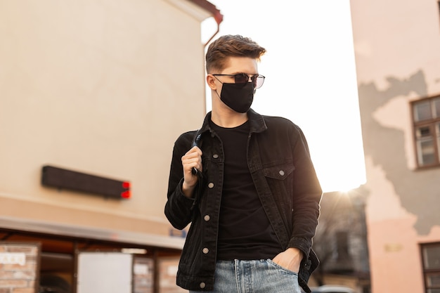 Giovane bell'uomo alla moda modello in elegante giacca di jeans nera con zaino in occhiali da sole vintage in maschera protettiva nera alla moda