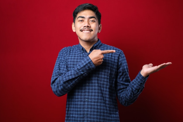 Giovane bell'uomo asiatico che indossa una camicia di flanella casual su sfondo rosso stupito e sorridente alla telecamera mentre si presenta con la mano e indica con il dito.
