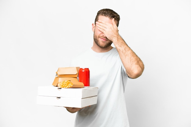 Giovane bell'uomo biondo che tiene pizze e hamburger su sfondo isolato che copre gli occhi con le mani Non voglio vedere qualcosa