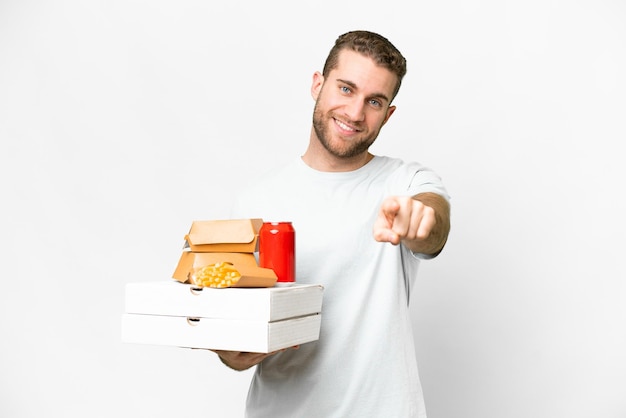 Giovane bell'uomo biondo che tiene pizze e hamburger su sfondo isolato che punta davanti con felice espressione