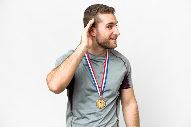 Giovane bell'uomo biondo con medaglie su sfondo bianco isolato ascoltando qualcosa mettendo la mano sull'orecchio
