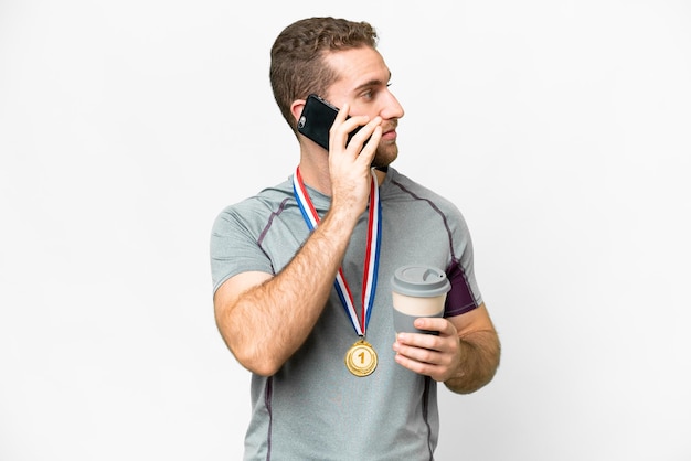 Giovane bell'uomo biondo con medaglie su sfondo bianco isolato che tiene il caffè da portare via e un cellulare