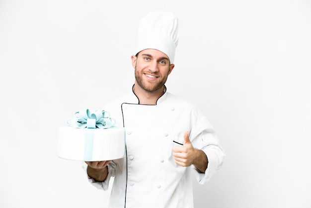 Giovane bell'uomo biondo con una grande torta su sfondo bianco isolato con i pollici in su perché è successo qualcosa di buono