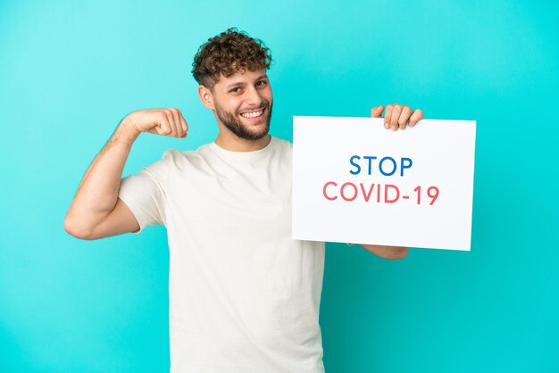 Giovane bell'uomo caucasico isolato su sfondo blu che tiene in mano un cartello con il testo Stop Covid 19 e fa un gesto forte