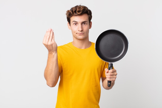 Giovane bell'uomo che fa un gesto di denaro o di denaro, dicendoti di pagare e tenendo in mano una padella da cucina