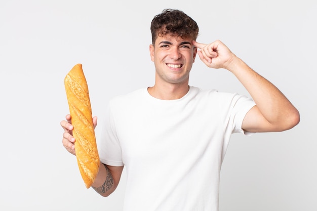 Giovane bell'uomo che si sente confuso e perplesso, mostra che sei pazzo e tiene in mano una baguette di pane