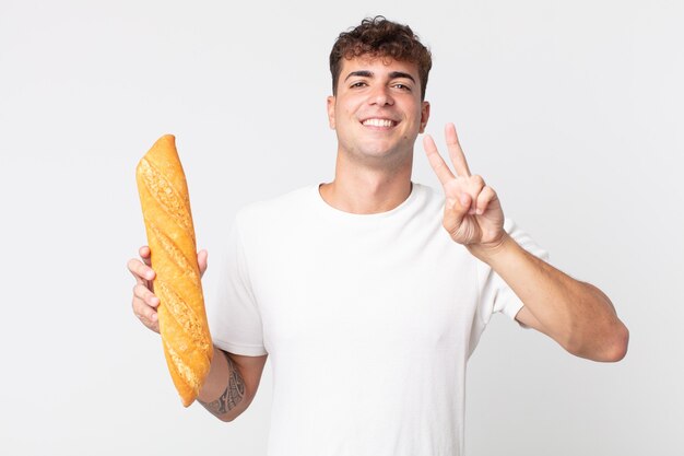 Giovane bell'uomo che sorride e sembra amichevole, mostra il numero due e tiene in mano una baguette di pane