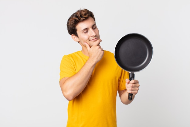 giovane bell'uomo che sorride felice e sogna ad occhi aperti o dubita e tiene in mano una padella da cucina