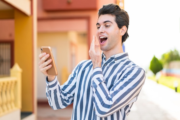 Giovane bell'uomo che utilizza il telefono cellulare all'aperto con espressione facciale sorpresa e scioccata