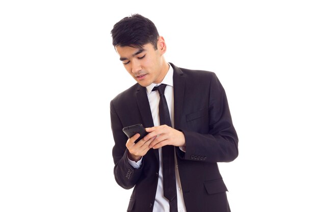 Giovane bell'uomo con i capelli neri in camicia bianca e abito nero con cravatta usando il suo telefono su sfondo bianco