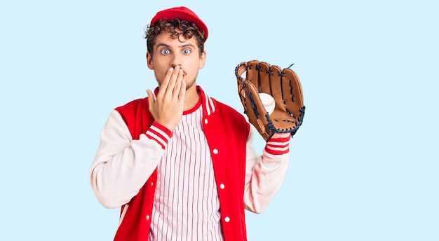 Giovane bell'uomo con i capelli ricci che indossa un'uniforme da baseball che tiene golve e palla che copre la bocca con la mano scioccata e impaurita per l'espressione sorpresa per errore