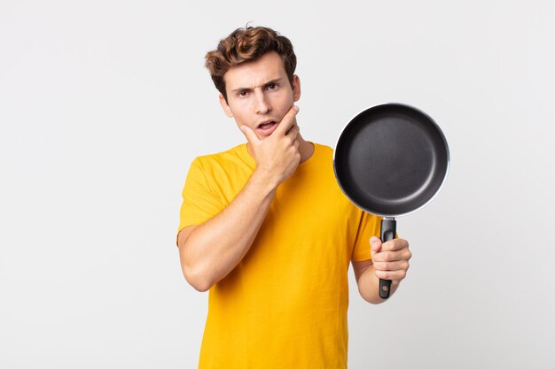 Giovane bell'uomo con la bocca e gli occhi spalancati e la mano sul mento e con in mano una padella da cucina