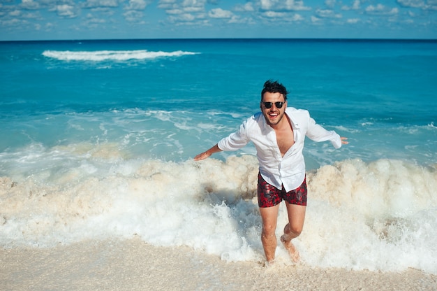 Giovane bell'uomo in camicia bianca e occhiali da sole che corre attraverso le onde del mare