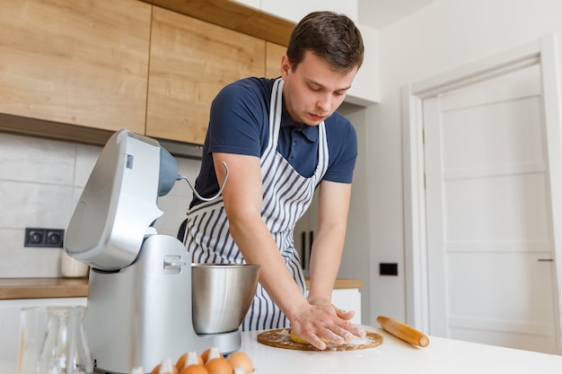 Giovane bell'uomo in grembiule che impasta la pasta in cucina Chef che fa pasticceria usando utensili a casa