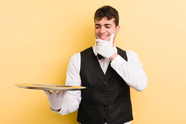 Giovane bell'uomo sorridente con un'espressione felice e sicura con la mano sul mento cameriere e concetto di vassoio