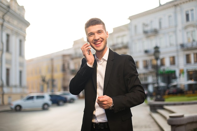 Giovane bell'uomo sorridente in giacca nera e camicia bianca che guarda felicemente da parte mentre parla al cellulare con bella vista sulla città su sfondo sfocato