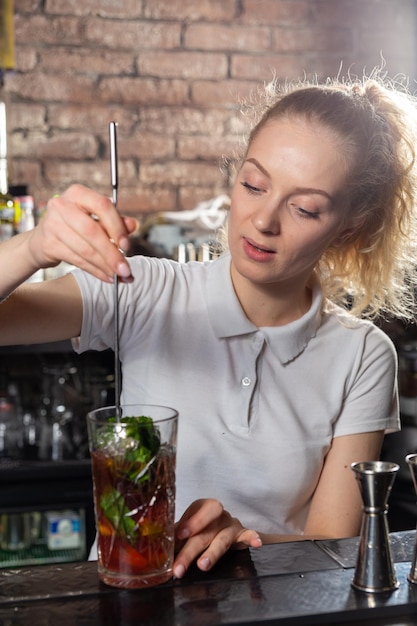 Giovane bella barista femminile che mescola un cocktail in un bicchiere alto