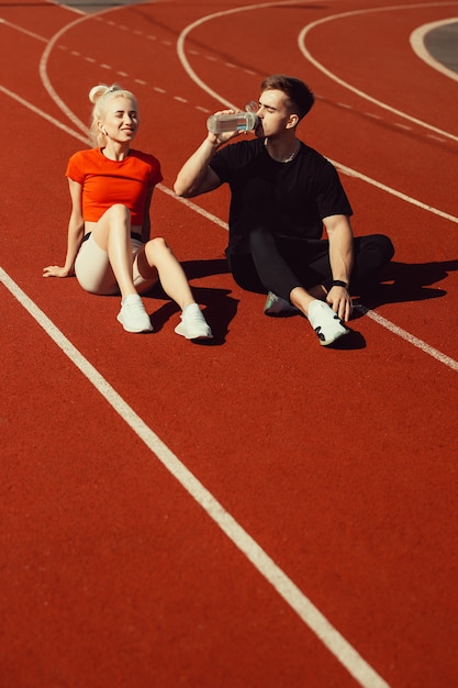 Giovane bella bionda e un ragazzo sono seduti su una pista da jogging con una bottiglia d'acqua e chiacchierano