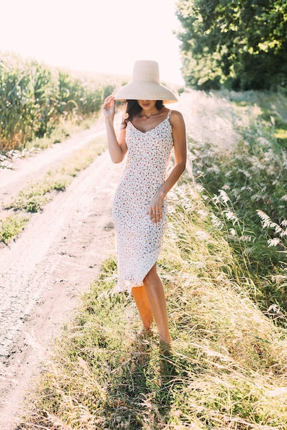 Giovane bella bruna in un elegante abito estivo e cappello di paglia sotto i raggi del sole sulla natura